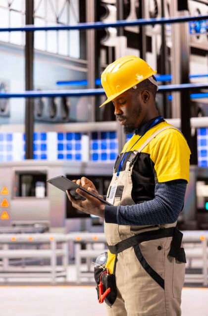 Male engineer analyzing industry 4.0 system performance in smart manufacturing plant. Black man monitoring machinery and robotic arms, ensuring optimal production and industrial automation efficiency.